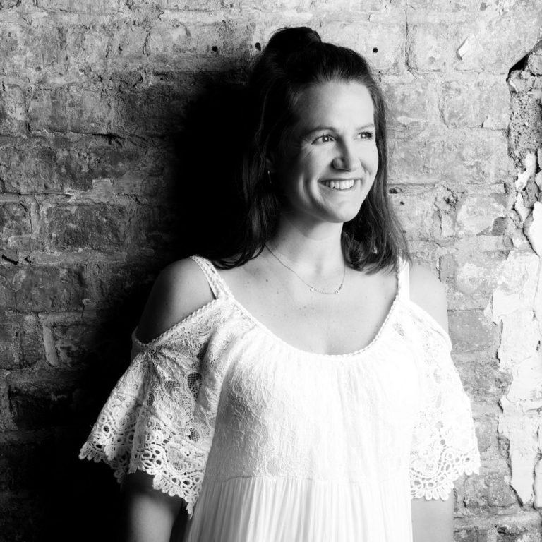 Ines Palms Hochzeitstanz Köln Frau in einem weißen, schulterfreien Kleid vor einer Backsteinwand.